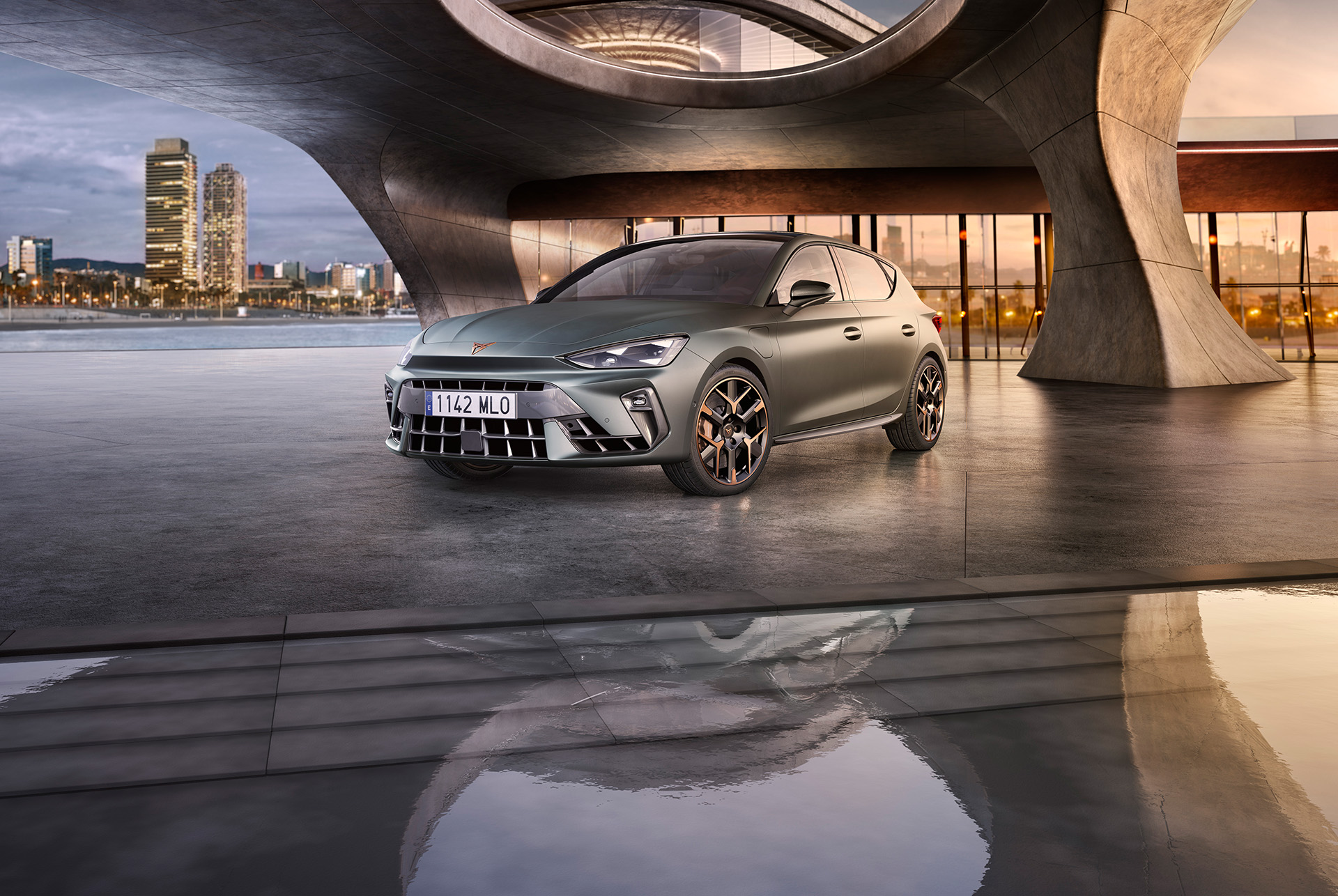 A new cupra leon 2024 in enceladus grey matte with copper accent alloy wheels parked on smooth concrete under an architectural structure. Barcelona cityscape in the background. 