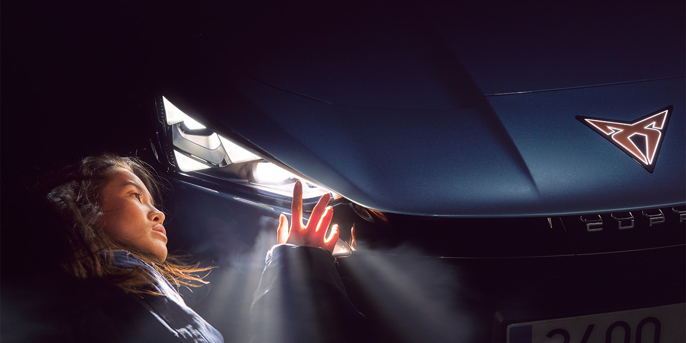 Close-up of a woman examining the CUPRA Tavascan's illuminated front logo at night.