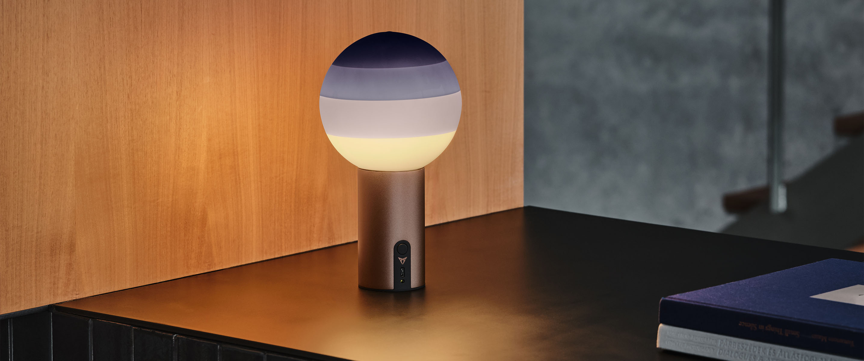 a portable cupra x marset dipping light with a spherical frosted glass top, purple gradient dip, emitting a soft glow and a cylindrical base against a wooden panel. the ambience is chic and contemporary. They feature metallic copper bases.