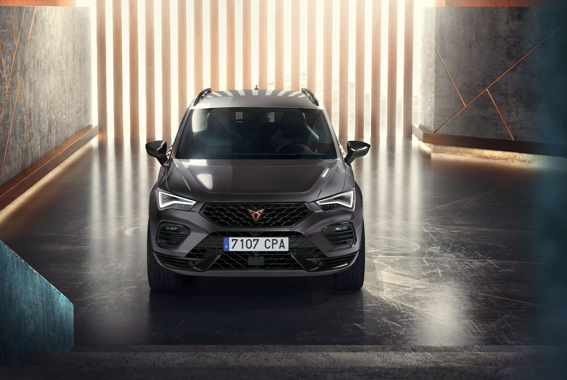 A front view of the Cupra Ateca, a grey SUV, in a modern, illuminated setting. The image highlights the car's bold front grille, LED headlights and sleek design. 