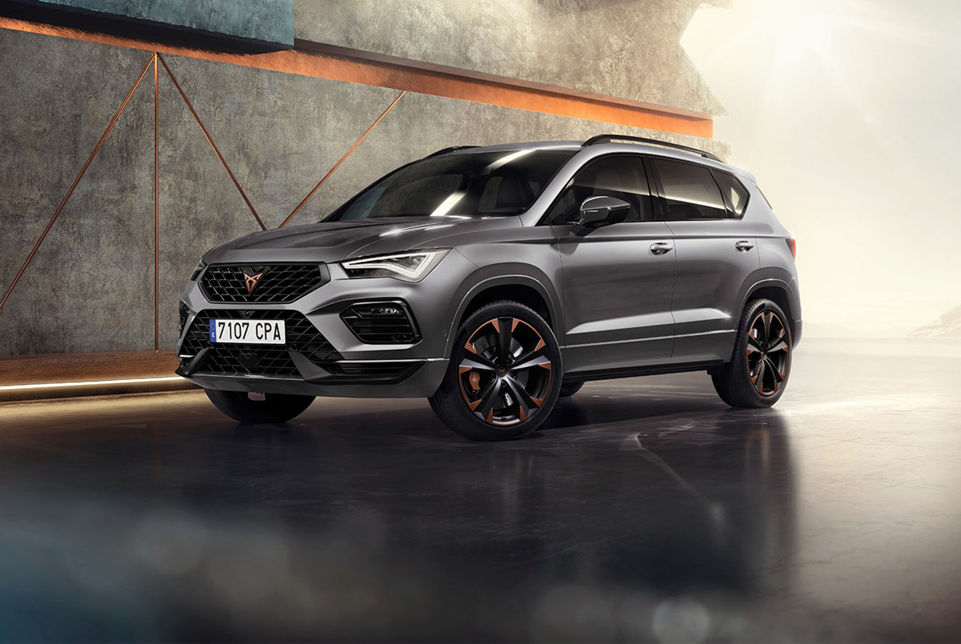 A 2024 cupra ateca in graphite grey, a sporty and compact suv parked in a modern, industrial setting. The car design features aerodynamic lines, led headlights and distinctive alloy wheels with copper accents. The background includes a geometric wall design, contrasting with the sleek vehicle design.