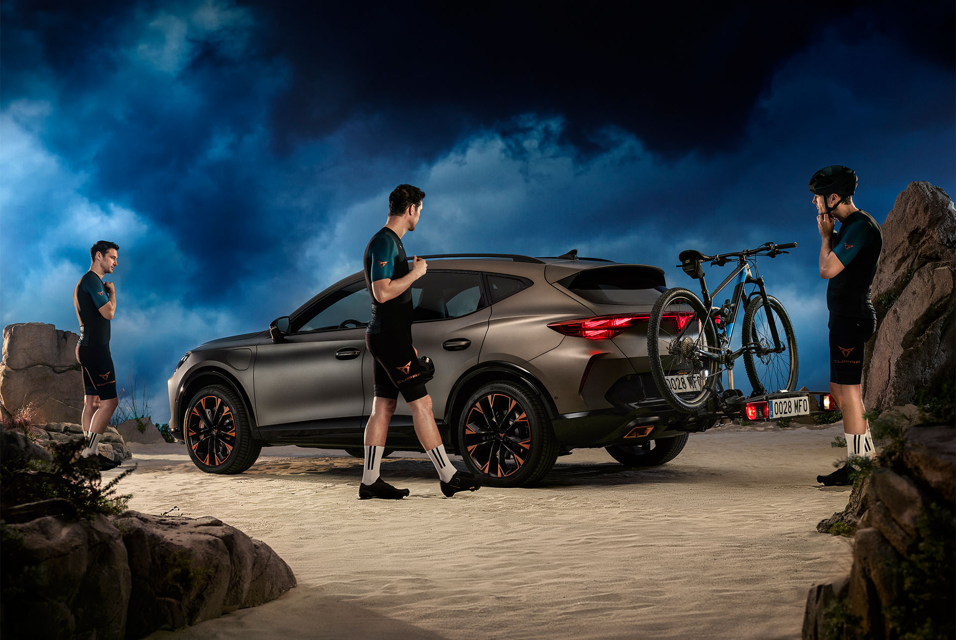 A CUPRA Formentor with a towing bike rack attached to the rear, holding two bicycles securely. The car is parked under a stormy sky, with cyclists standing around the vehicle, preparing for a ride.