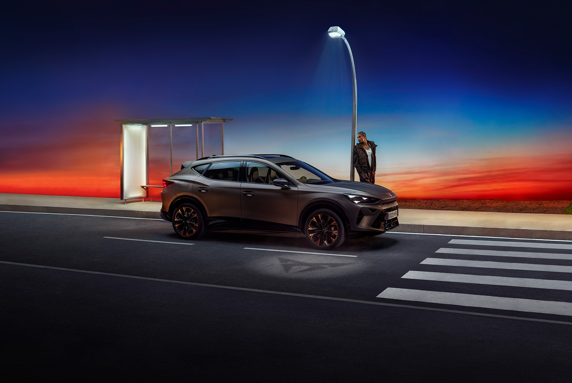 CUPRA Formentor in century bronze matt, parked in a building facing a red sky, being charged by an individual next to it.