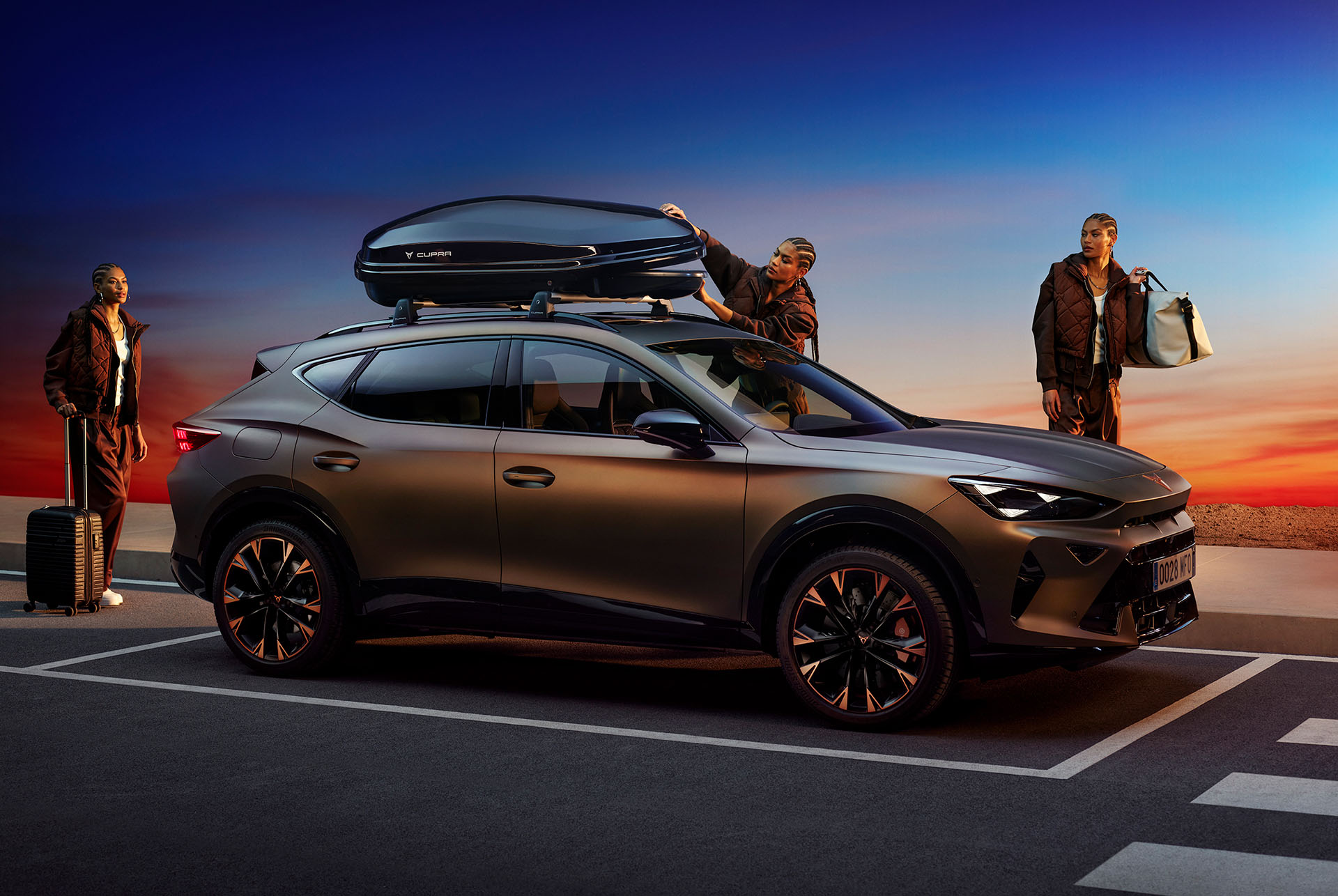 CUPRA Formentor with a sleek, aerodynamic roof box mounted on top, designed for extended journeys and extra gear storage, set against a scenic sunset backdrop with three individuals standing nearby.