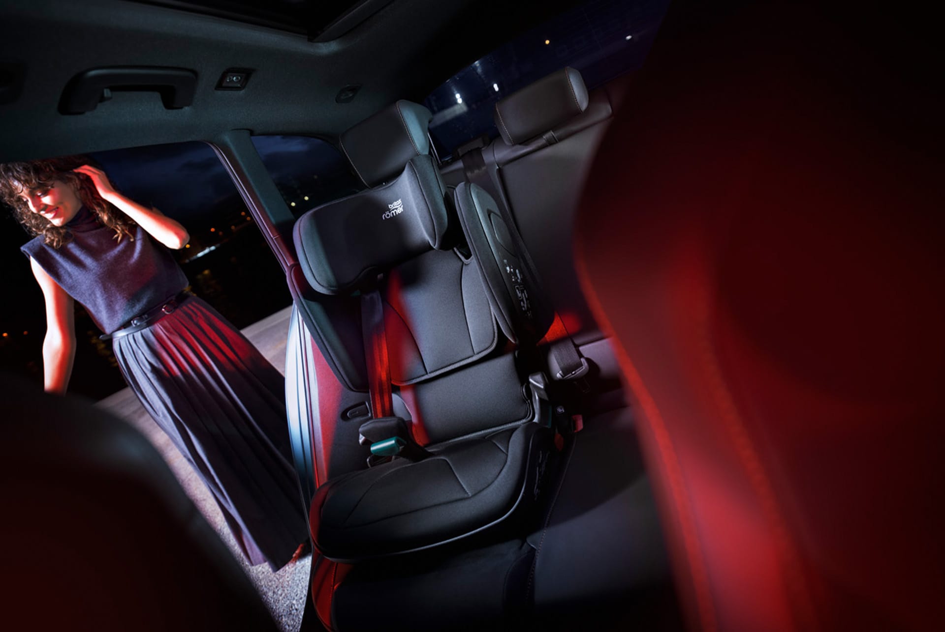 the interior design of a matte grey CUPRA Leon Sportstourer. The photo is focus in the child seat. A woman is standing outside the car, looking in. The car's interior is dark, with red lighting highlighting the seats and dashboard.