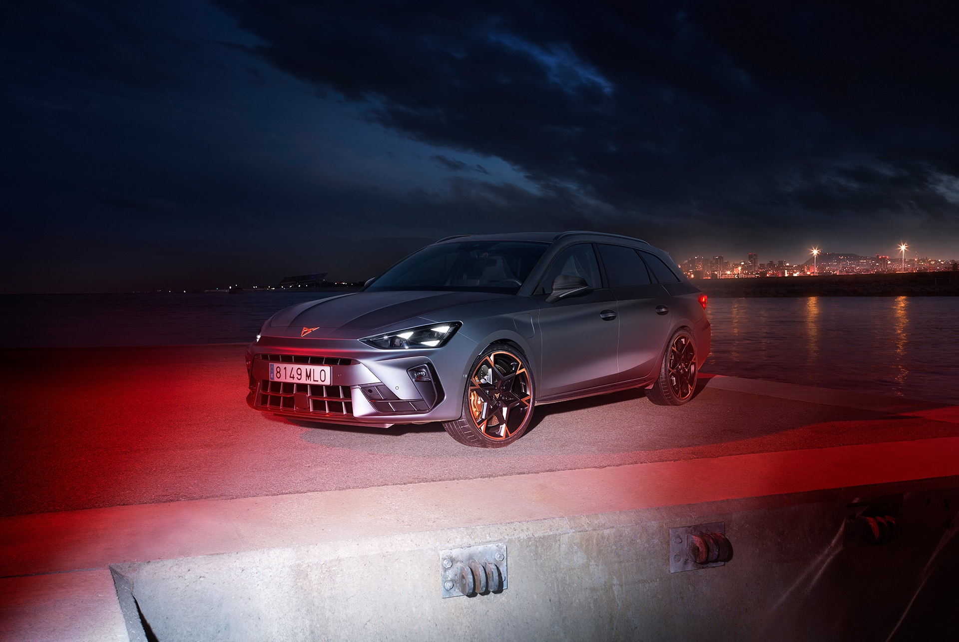 A CUPRA Leon Sportstourer 2024 in matte grey is parked on a waterfront at night. The city skyline is visible in the background, with its lights reflecting in the water. The car is illuminated by red lights, creating a dramatic and eye-catching scene.