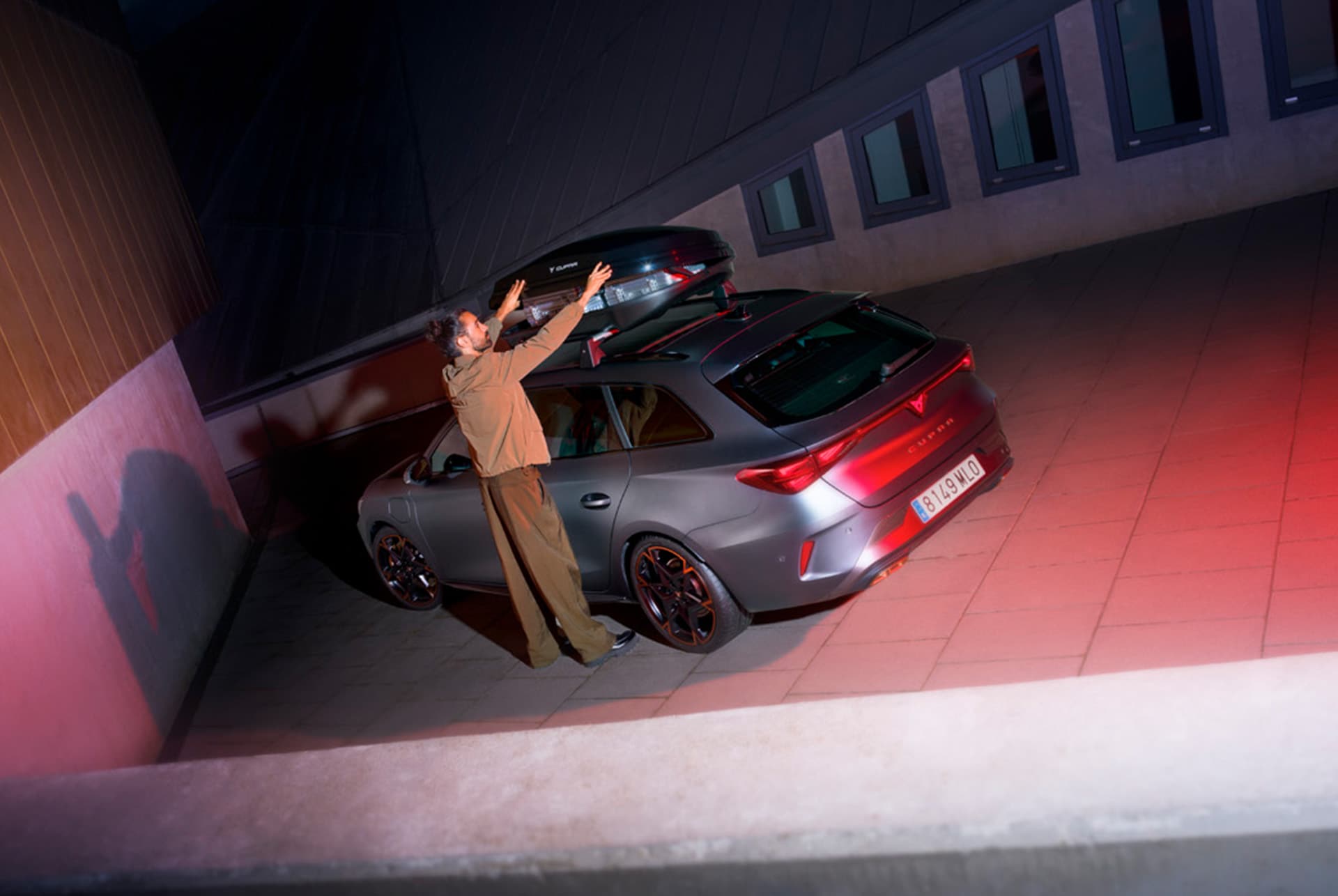 A man is standing next to a matte grey CUPRA Leon Sportstourer with a large roof box attached to it. The car is parked in a dimly lit garage or driveway. The scene is illuminated by red lights, creating a dramatic atmosphere.