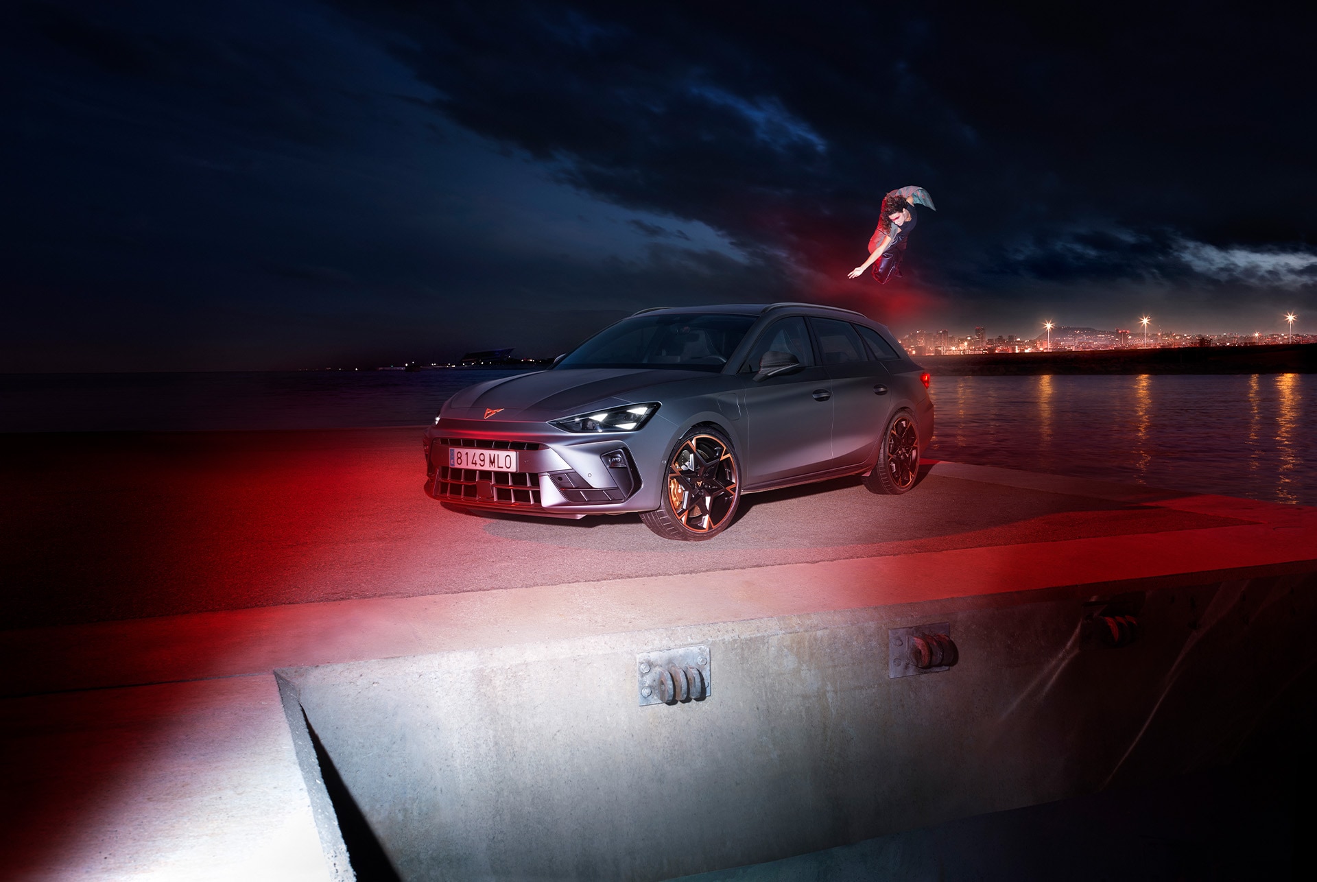 A side view of the CUPRA Leon Sportstourer in matte grey. The car is parked on a waterfront at night. It is illuminated by red lights, creating a dramatic contrast against the dark sky. the background, a person is performing a dynamic jump or flip, adding a sense of energy and movement to the scene. The city skyline is visible in the distance.