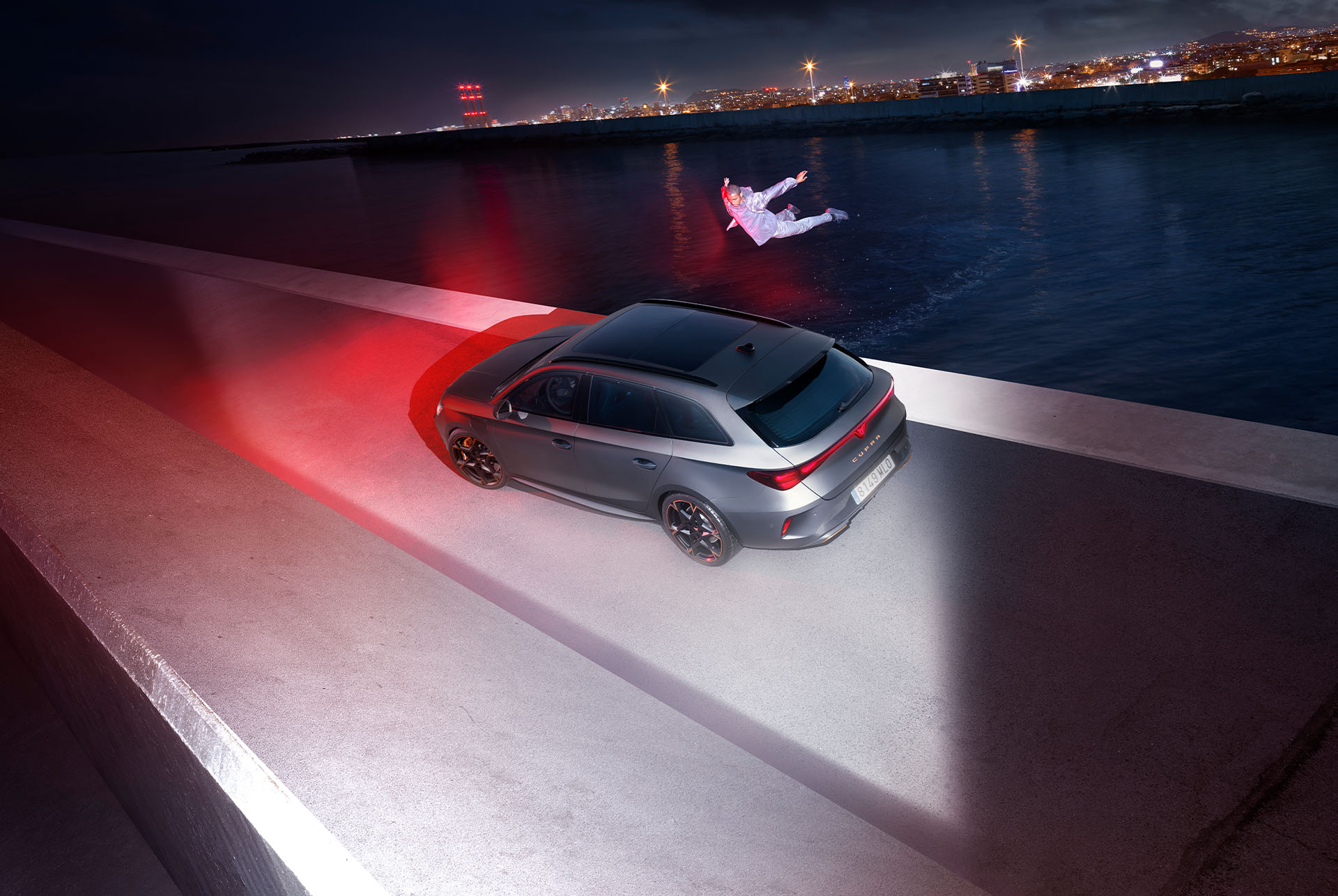 The image shows a dramatic scene with a matte grey CUPRA Leon Sportstourer parked near a body of water. In the foreground, a person is leaping into the water, creating a sense of motion and excitement. The car is illuminated by red lights, highlighting its sleek design and sporty stance.