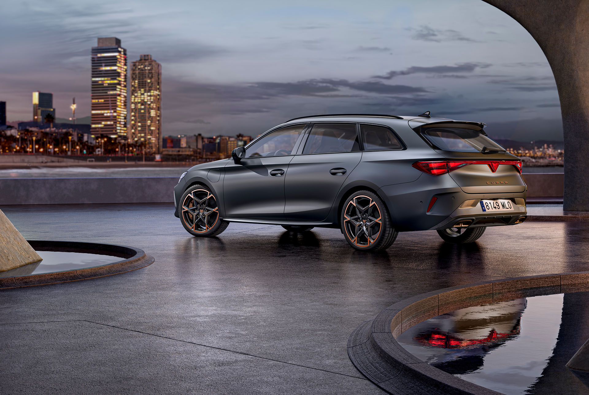 New cupra leon sportstourer 2024 technology parked on cement, structure with glowing edges, overlooking a city skyline at dusk. The vehicle is shown in a matte grey finish with copper and black alloy wheels.