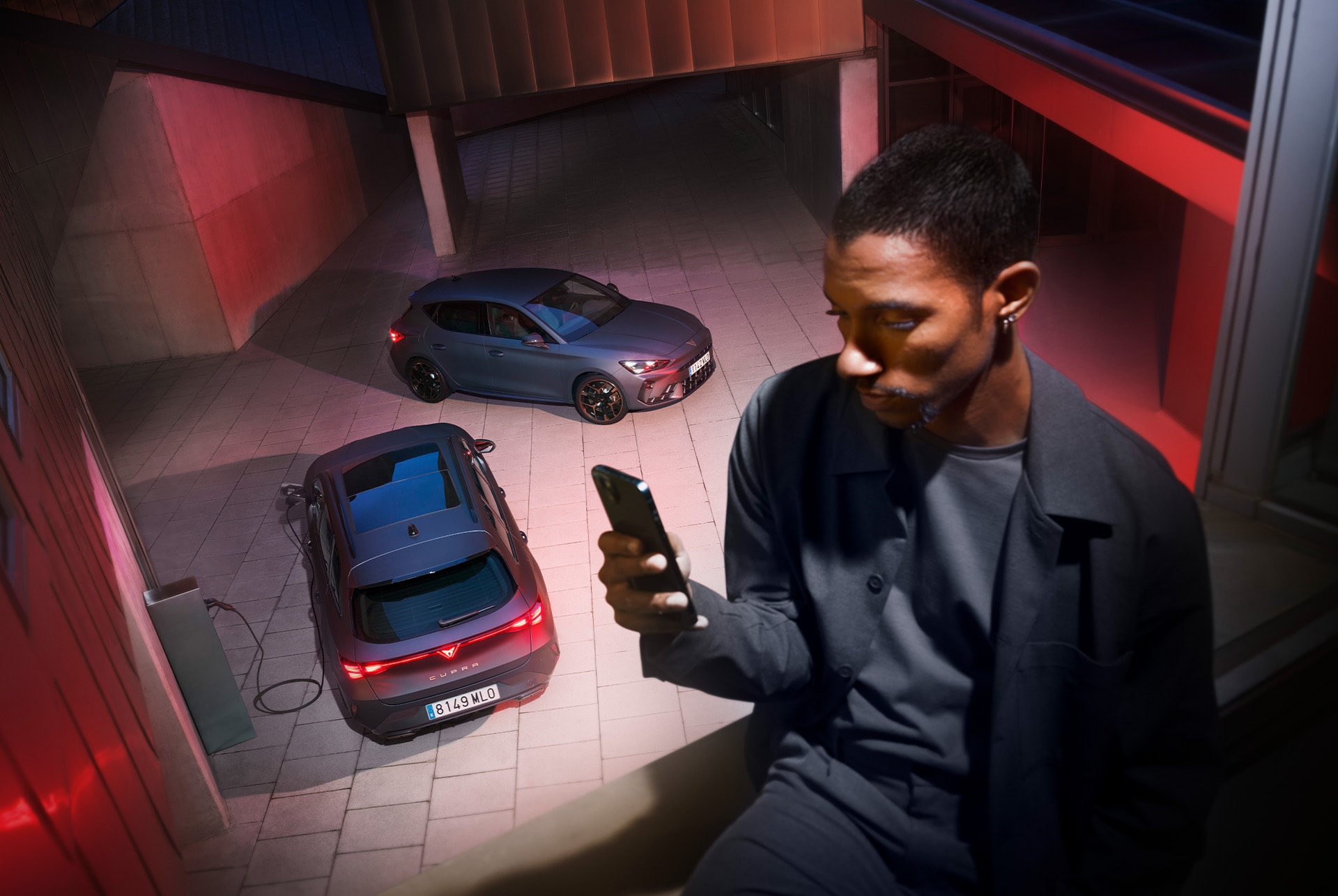 in the foreground the image of a man holding a telephone. On the phone, we sense, he was looking at the Curpa App. In the background, as if they were on the bottom floor of the building, we see 2 Cupra vehicles, one from the front view and the other from the rear view.