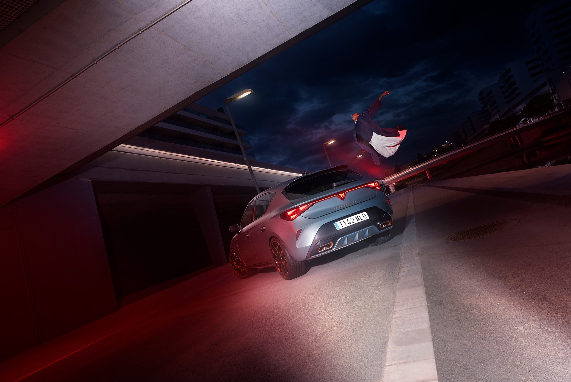 A CUPRA Leon is parked under an overpass at night. A person is jumping in the background, creating a sense of motion and excitement. The car is illuminated by red lights, highlighting its sporty design and aggressive rear end.