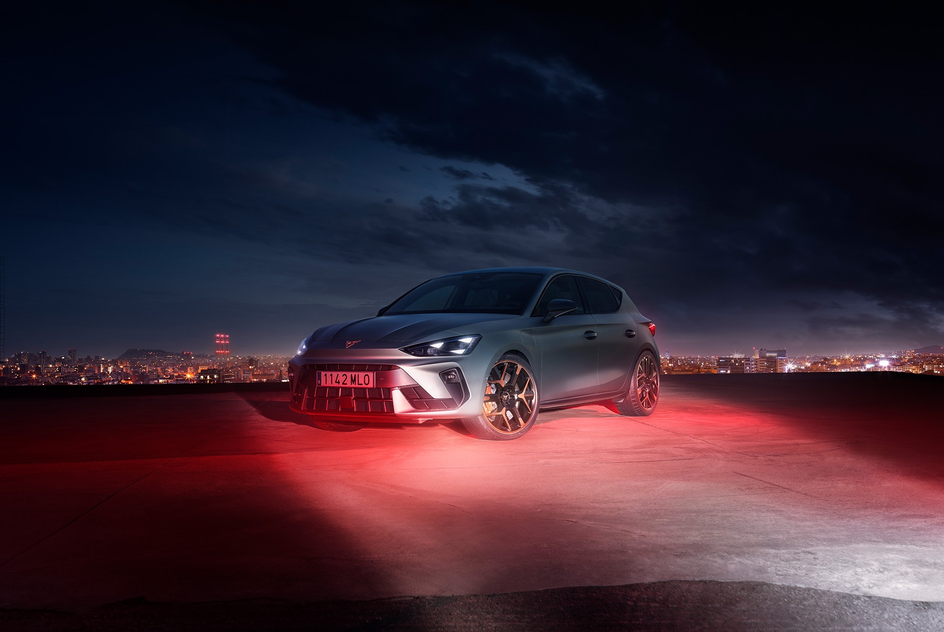 A sleek, matte grey CUPRA Leon is parked on a rooftop at night. The city skyline is visible in the background, with its lights creating a vibrant glow. The car is illuminated by red lights, highlighting its sporty design and aggressive front end.