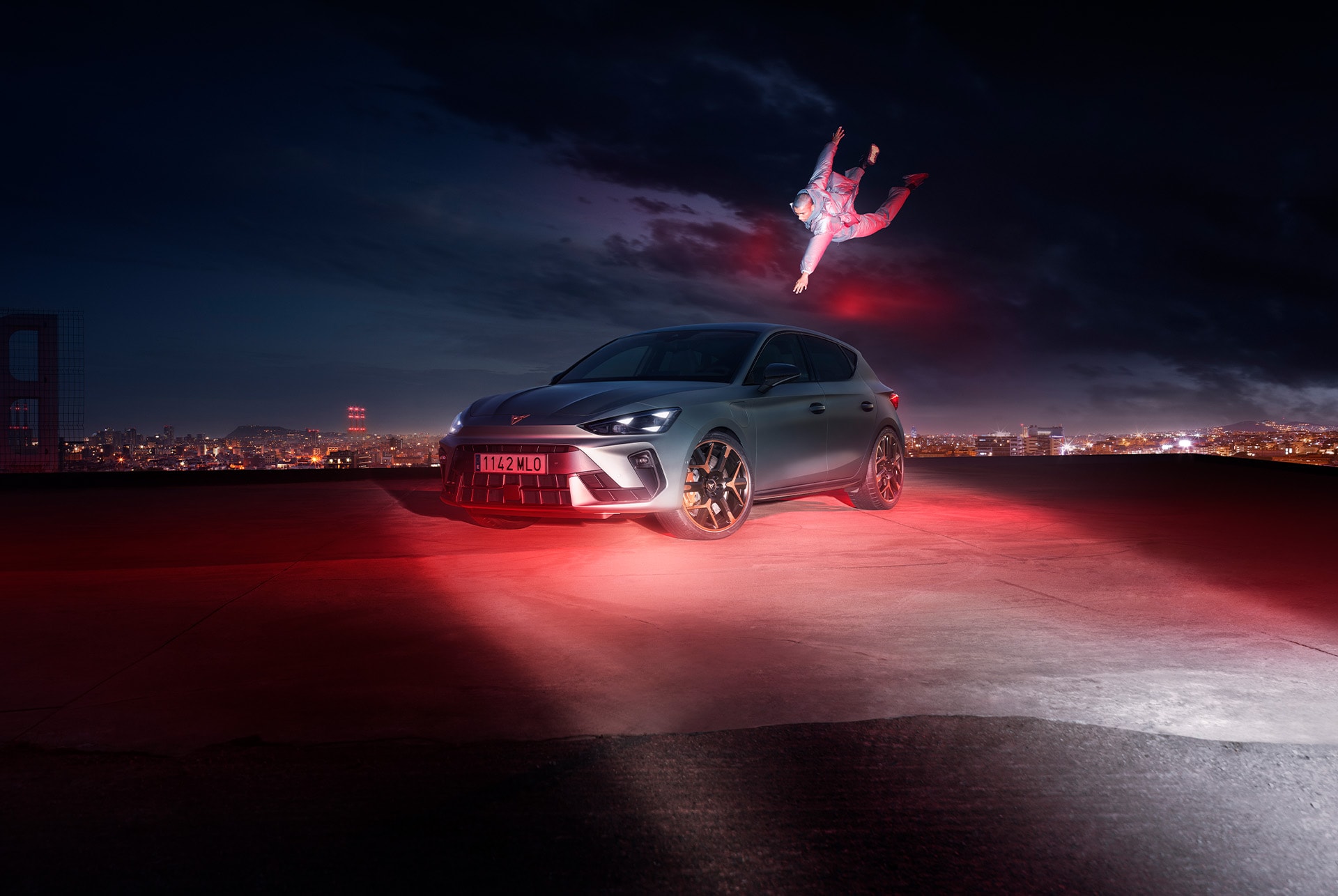 CUPRA Leon in matte grey is parked on a rooftop at night. A person is performing a dynamic jump in front of the car, adding a sense of thrill to the scene. The city skyline is visible in the background, with its lights creating a vibrant glow. The car is illuminated by red lights, highlighting its sporty design and aggressive front end