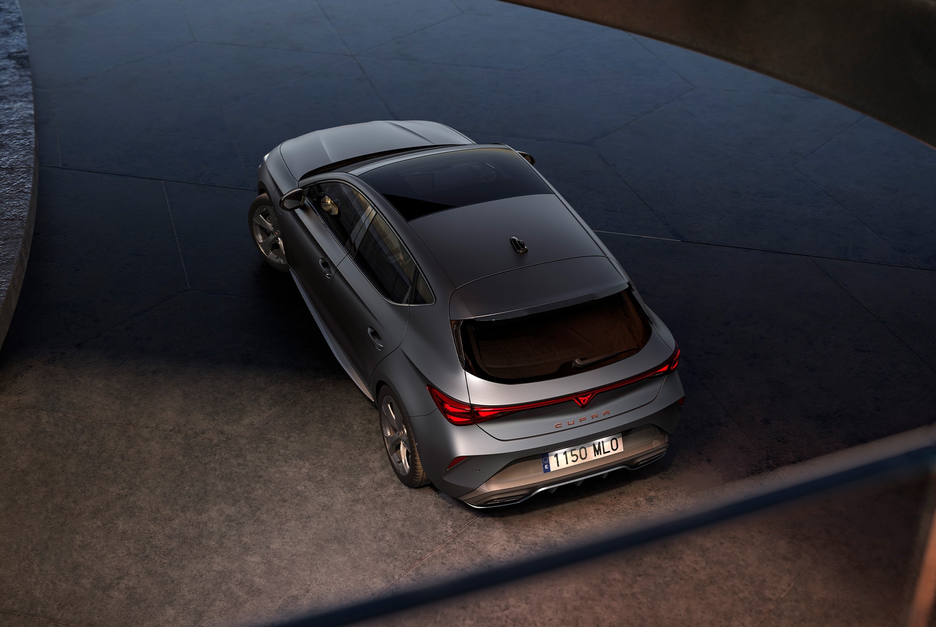 A top-down view of a sleek, matte grey CUPRA Leon. The car is parked on a dark surface. The car has a panoramic sunroof and distinctive alloy wheels. 