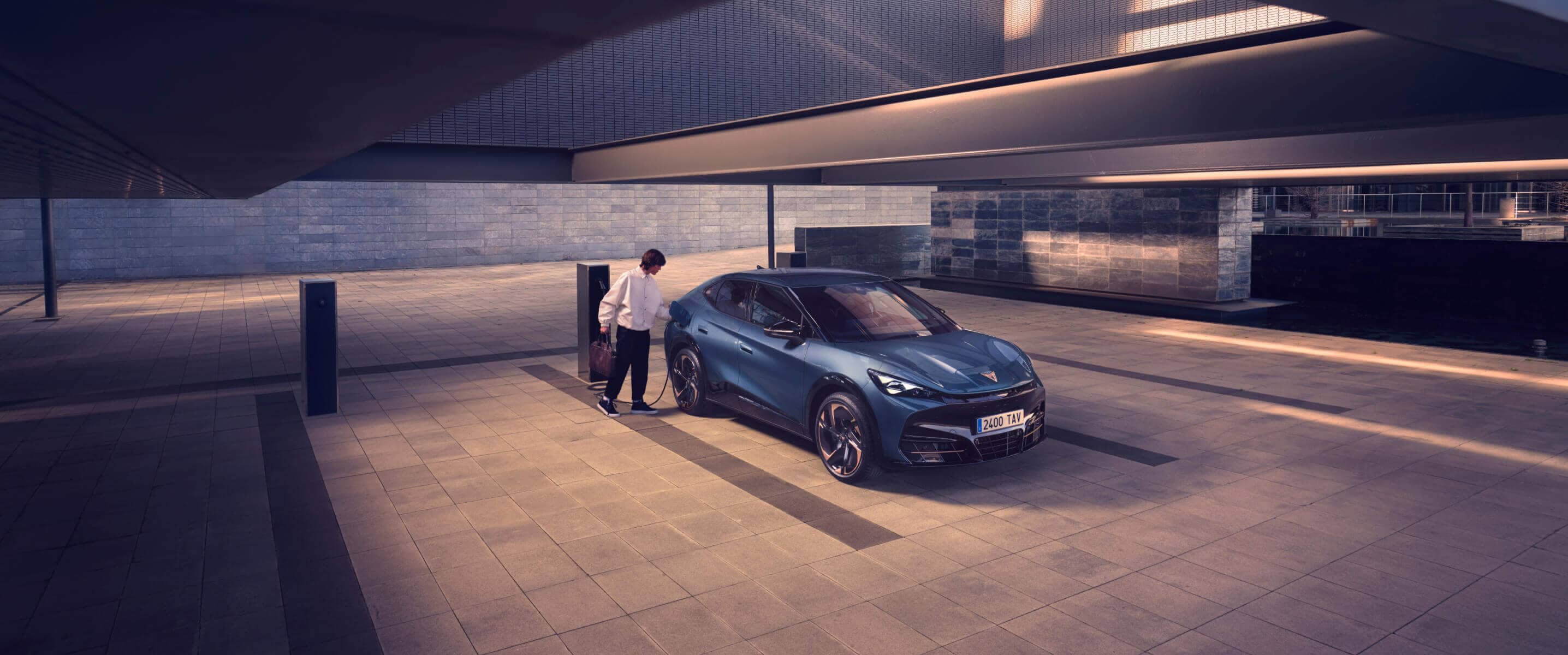 A bright blue CUPRA charging in an internal parking and a woman dressed in white next to the car.