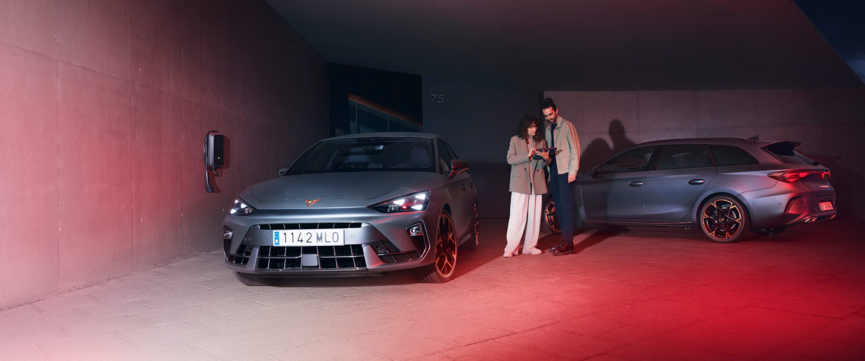 A dark grey CUPRA electric vehicle, being charged next to a EV Charger with a sunset as the background.