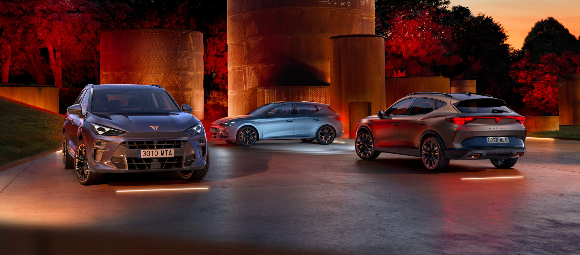 Three plug in hybrid vehicles, parked outside in a park.With a dimmed light that surrounds them and trees in the background.