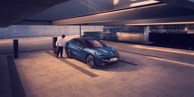 A bright blue CUPRA charging in an internal parking and a woman dressed in white next to the car.