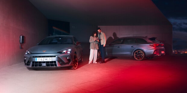 A dark grey CUPRA electric vehicle, being charged next to a EV Charger with a sunset as the background.